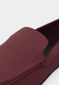 Κοντινό πλάνο ενός μπορντό παπουτσιού slip-on με λείο υλικό και ορατή ραφή κατά μήκος του επάνω μέρους και της πλευρικής ραφής.