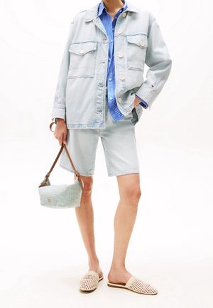 Personne portant une veste en denim clair et un short par-dessus une chemise rayée bleue, tenant un petit sac à main rayé, portant des chaussures beiges tissées sans lacets.