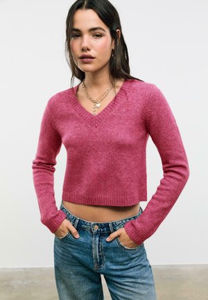 Mlada žena s dugom tamnom kosom, u ružičastom skraćenom V-izrez sv sweateru, s višeslojnim zlatnim ogrlicama i plavim trapericama, s rukama u džepovima.