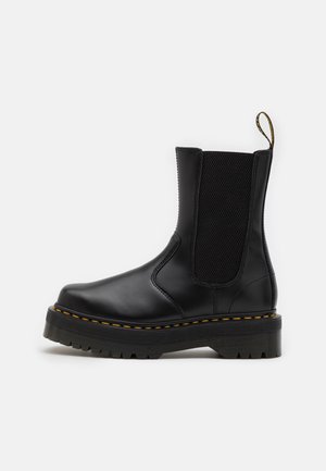 Botte Chelsea en cuir noir avec panneau latéral élastique, coutures jaunes sur une semelle épaisse à crampons, et languette de traction à l'arrière.