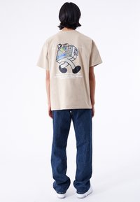 Camiseta gráfica beige con un diseño colorido de maleta en la espalda, combinada con unos jeans de denim azules de corte holgado y zapatillas blancas.