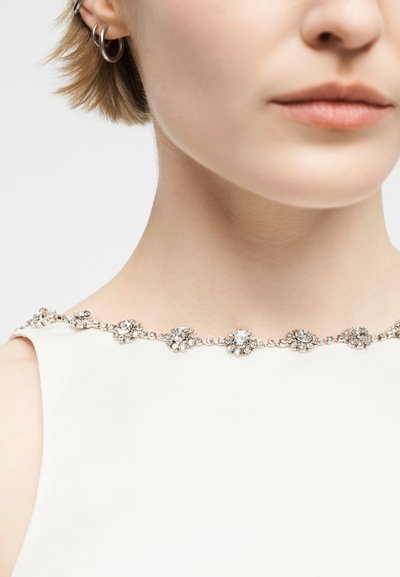 Femme portant un haut blanc sans manches avec des ornements floraux en strass le long de l'encolure et des boucles d'oreilles argentées en forme d'anneau.