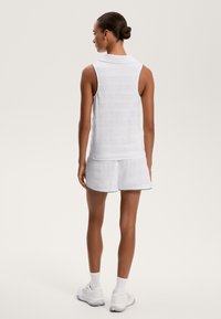 Bela brezrokavnica s polo ovratnikom z teksturiranimi črtami, kombinirana z belimi kratkimi hlačami, ki imajo črn rob. Obuta v bele športne čevlje.