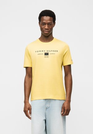 Młody mężczyzna w żółtym T-shircie Tommy Hilfiger z czarnym logo, jasnoniebieskie dżinsy, stojący na tle jednolitej białej ściany.