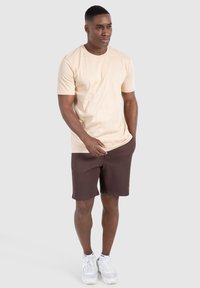 Lys beige bomulds t-shirt og brune shorts, der har en afslappet pasform og korte ærmer. Modellen står med hænderne i lommerne og har hvide sneakers på.