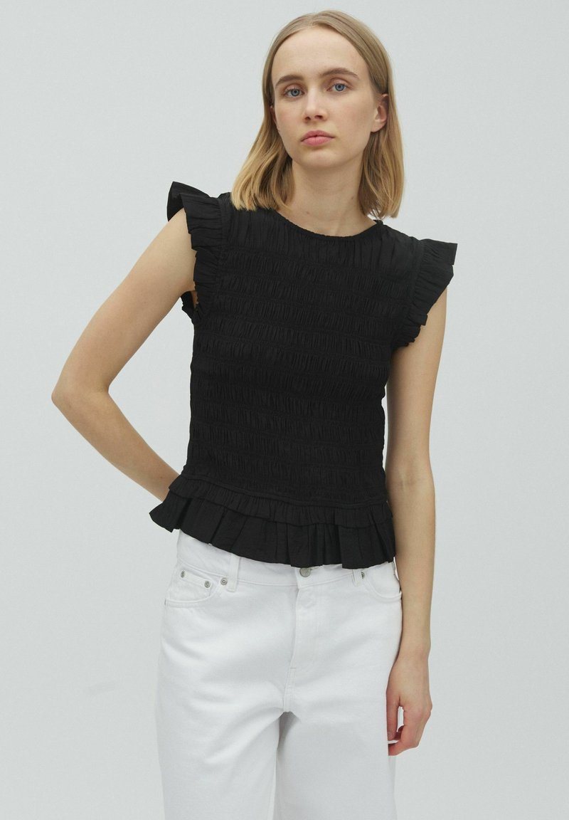 Jeune femme aux cheveux blonds et raides portant un haut noir sans manches à volants et un pantalon blanc taille haute, debout devant un fond uni.