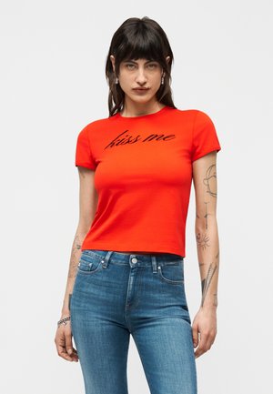 Femeie cu păr închis la culoare, purtând un tricou roșu scurt cu textul „sărută-mă” și blugi albaștri, arătând mai multe tatuaje pe brațe pe un fundal simplu.