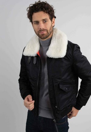 Veste bomber noire avec un col en fausse fourrure blanche, fermeture éclair à l'avant, poches latérales et poignets côtelés. Portée sur un pull gris.