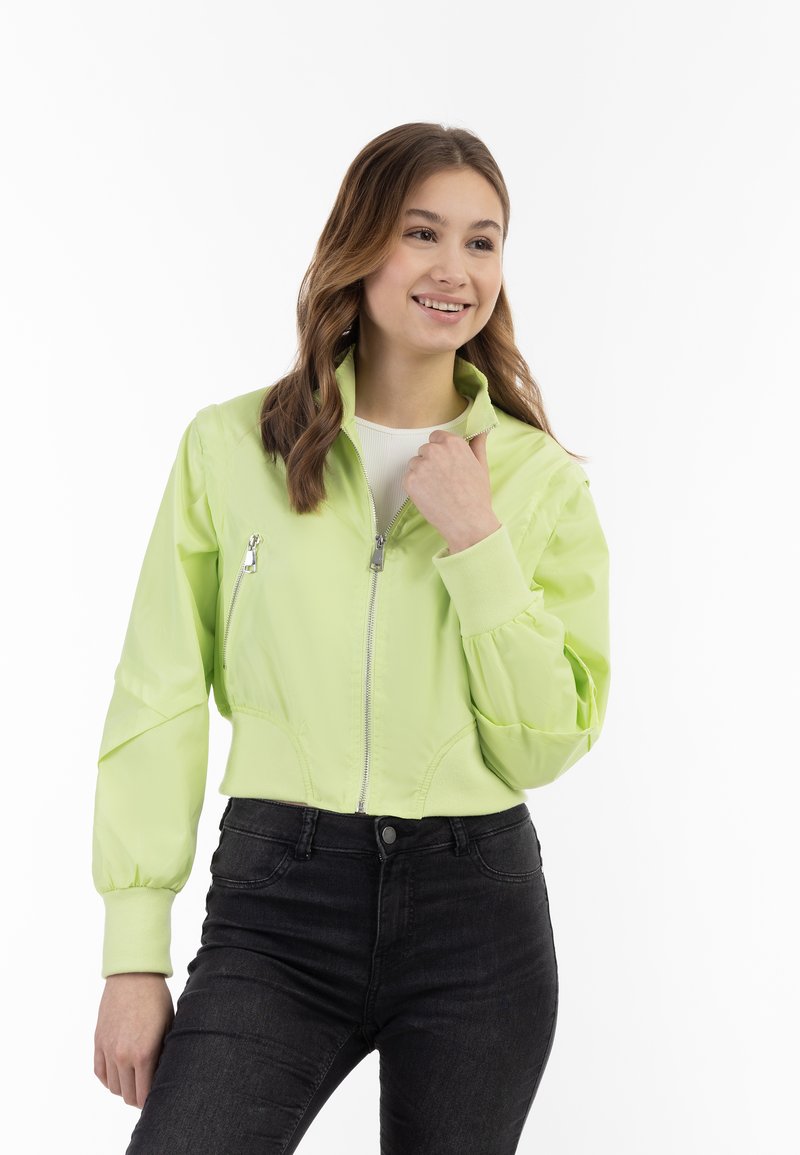 Giacca bomber cropped verde chiaro con zip frontale, tasche laterali e polsini elasticizzati, abbinata a jeans neri. Texture liscia.