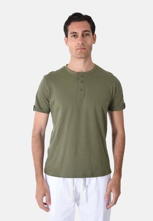 Uomo in piedi rivolto in avanti, indossa una maglietta henley verde oliva a maniche corte e pantaloni bianchi su uno sfondo semplice.