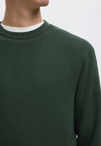 Person iført en mørkegrøn crewneck-sweatshirt over en hvid skjorte, med kun nakke og skulder synlige, vendt væk.