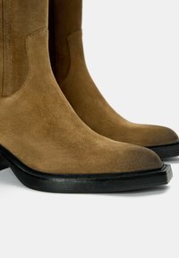 Bottes en suede marron avec semelles noires, présentant un design à bout carré et une texture lisse, présentées en gros plan sur fond blanc.