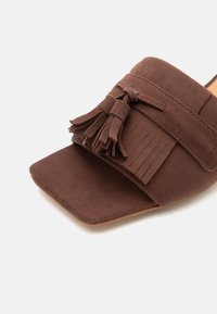 Καφέ μουλάρια από σουέτ με τετράγωνη μύτη, με διακοσμητικές φούντες και φραγκοράμματα σε ανοιχτό φόντο.