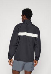 Water-resistant black jacket with a high collar and a white horizontal stripe across the back. Features long sleeves and lightweight material.