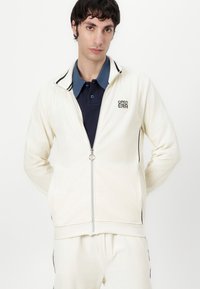 Weißes, geripptes Zip-Jacke mit schwarzen Akzenten, ausgestattet mit einem Stehkragen und einem Logo. Über einem marineblauen Poloshirt getragen.