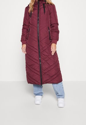 Femme portant un long manteau d'hiver matelassé bordeaux avec une fermeture éclair à l'avant, des cordons noirs, un jean bleu et des baskets blanches.