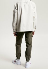 Vaalean beige paita, vihreät cargo-housut joustavilla resoreilla ja valkoiset lenkkarit neutraalilla taustalla. Yksinkertainen muotoilu hienovaraisilla yksityiskohdilla.