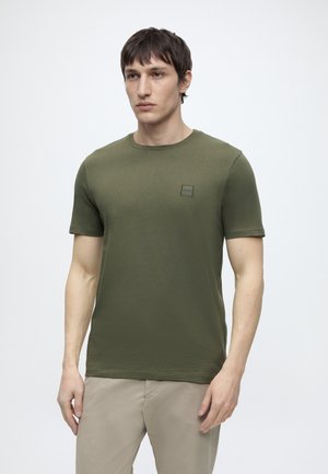 Fiatal férfi, aki olíva zöld rövid ujjú pólót és bézs nadrágot visel, egy egyszerű világos háttér előtt áll.