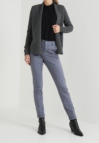 Grau gerippter Cardigan über einem schwarzen Hemd mit Knöpfen, kombiniert mit hellgrauen Slim-Fit-Hosen und schwarzen Ankle-Boots.
