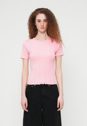 Junge Frau mit lockigem rotem Haar, die ein rosa, kurzärmeliges, geripptes Oberteil und schwarze, weit geschnittene Hosen trägt, steht vor einem einfarbigen weißen Hintergrund.