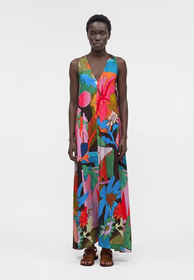 Femme portant une robe longue sans manches à col en V avec un motif floral et feuillage multicolore et éclatant, accompagnée de sandales marron.