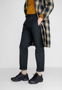 Černé sportovní boty, tmavé kalhoty s přizpůsobeným střihem a hořčicová košile s leopardím vzorem pod károvaným kabátem v béžových a námořnických tónech.