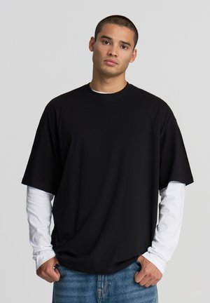 T-shirt nera oversize con maniche corte sovrapposta a una camicia bianca con maniche lunghe. Realizzata in cotone, presenta un collo rotondo e una vestibilità comoda.