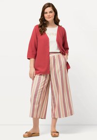 Koralli neulejakku, jossa on kolme neljännes hihaa, valkoisen topin päällä, yhdistetty leveisiin, vaaleanpunaisiin ja beigeisiin raitahousuihin. Ruskeat sandaalit.