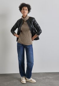 Černá kožená bomber bunda, béžový pletený rolák, modré džíny a béžové tenisky. Kudrnaté vlasy, stojící póza v neutrálním prostředí.