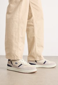 Beige suède sneakers met textuurpanelen, donkerblauwe accenten en witte veters. Rubberen zool met een contrasterend rood logo op de hiel.