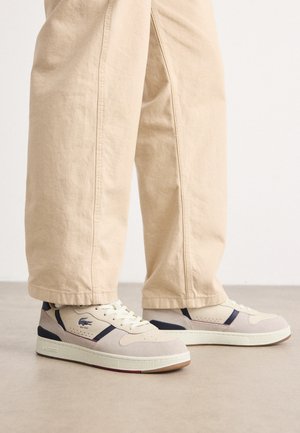 Beżowe zamszowe sneakersy z teksturowanymi panelami, ciemnoniebieskimi akcentami i białymi sznurowadłami. Gumowa podeszwa z kontrastującym czerwonym logo na pięcie.