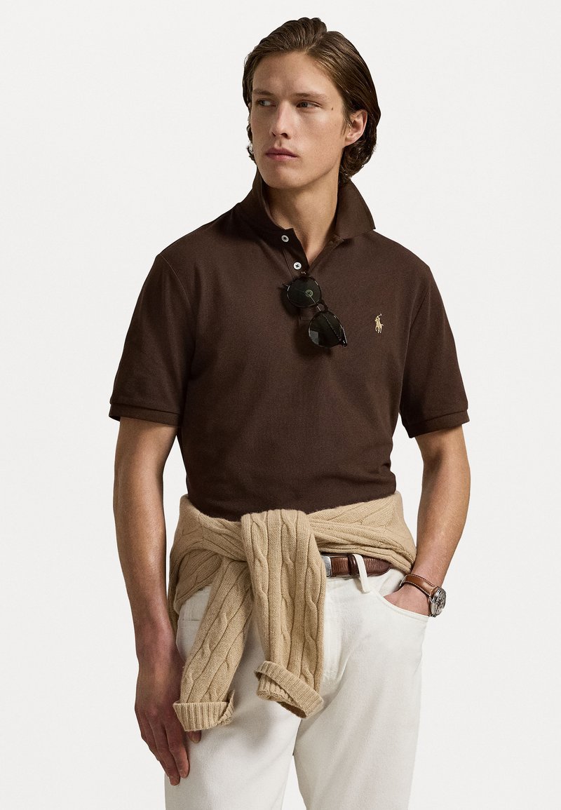 Polo shirt marron à manches courtes avec col, doté de détails de boutons et d'un petit logo. Pull en maille câblée beige clair noué autour de la taille.
