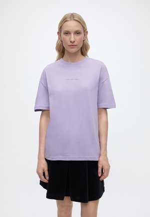 Donna bionda in piedi, che indossa una camicia oversize a maniche corte color lavanda con un piccolo testo sul petto e una gonna nera a pieghe.