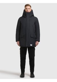 Czarna pikowana kurtka z wysokim kołnierzem i zamkiem błyskawicznym, z dwoma przednimi kieszeniami. Noszona na czarnych spodniach typu cargo i butach.