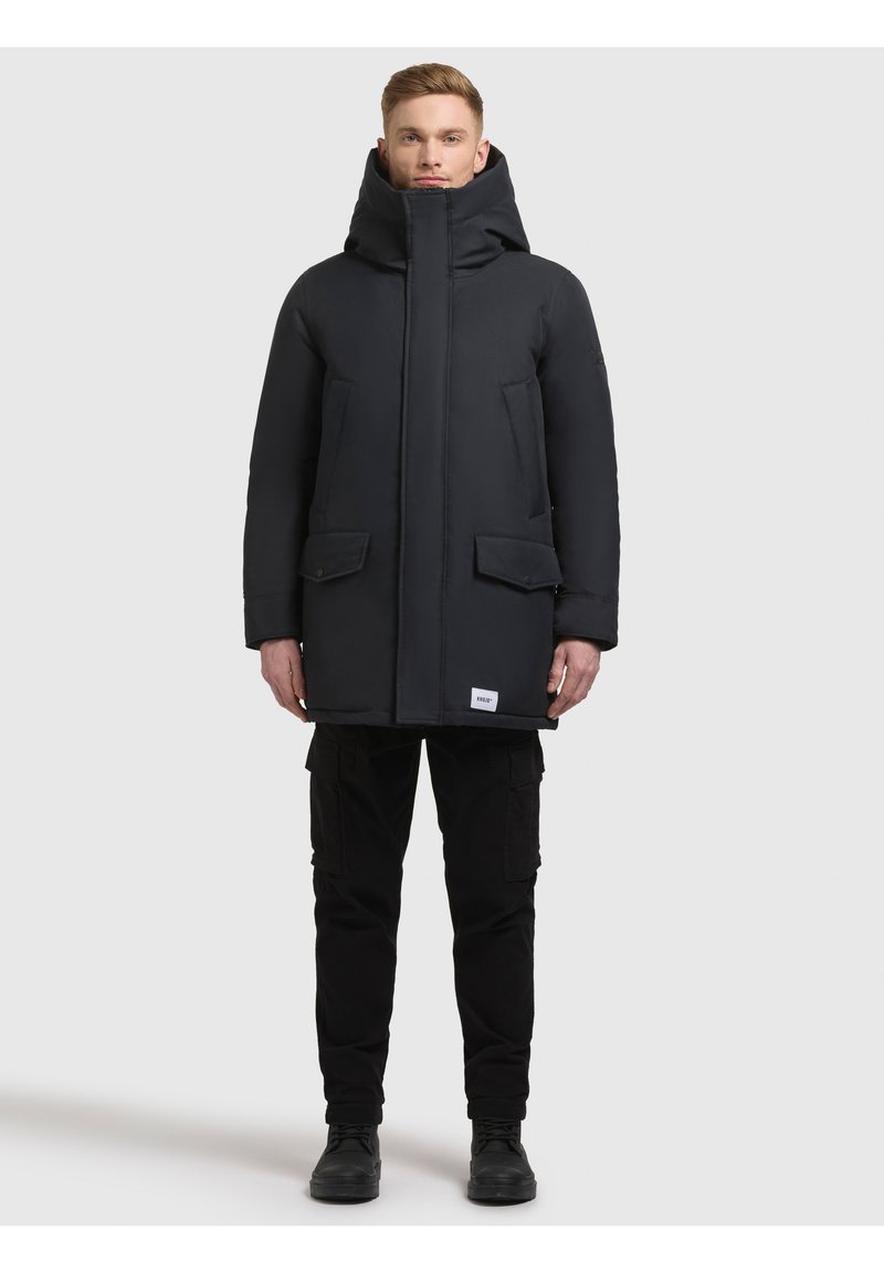 Czarna pikowana kurtka z wysokim kołnierzem i zamkiem błyskawicznym, z dwoma przednimi kieszeniami. Noszona na czarnych spodniach typu cargo i butach.