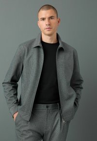 Chaqueta gris con patrón, cuello estructurado, cierre de cremallera y mangas largas, combinada con una camisa negra ajustada y pantalones grises a juego.