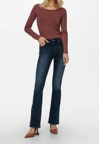 Jean flare en denim bleu foncé avec des détails de délavage subtils, associés à un haut fitted à manches longues bordeaux et des chaussures à talons hauts marron.