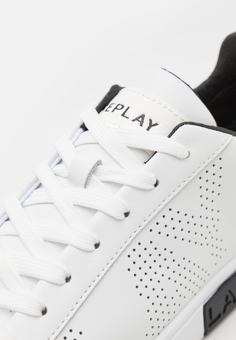 Weißer Sneaker mit perforierter Textur, flachen weißen Schnürsenkeln und schwarzen Akzenten an der Ferse und im Logo. Belastbares Material und minimalistisches Design.