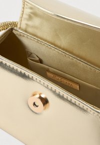 Fémes arany táska sima felülettel, belső cipzáras zsebbel és márka címkével, kerek gombbal zárva.