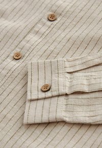 Beige stof skjorteærme og forstykke med tynde lodrette striber og runde brune knapper.