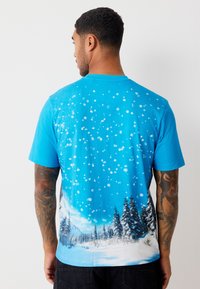 Modré tričko s krátkým rukávem s potiskem zasněžené krajiny na zádech, s bílými sněhovými vločkami a stálezelenými stromy na spodní části.