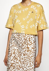 Gul bomullströja med vit text och Jordan-logga, matchad med en vit kjol med bruna prickar. Korta ärmar, avslappnad passform.