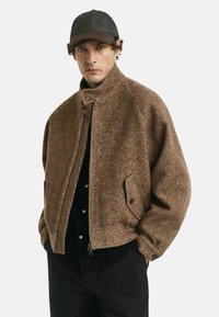 Bruine visgraatjas met een hoge kraag en knoopsluiting, voorzien van zijzakken. Draagt over een zwart shirt, wat past bij donkere broeken.