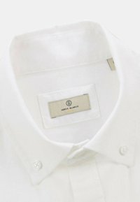 Chemise blanche à col, réalisée en coton, avec un design à boutons, une texture subtile et une étiquette avec le nom de la marque cousue à l'intérieur du col.