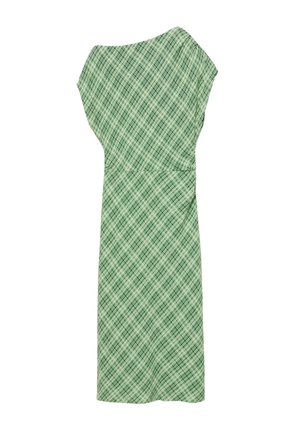 Jeune femme portant un haut à manches courtes à carreaux verts avec une épaule découverte, les bras croisés, regardant pensivement sur le côté sur un fond uni.