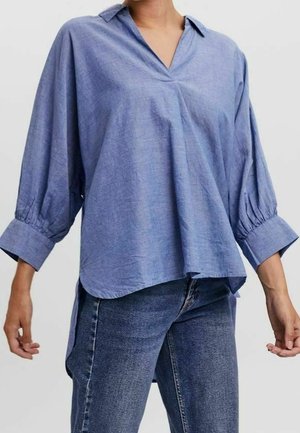 Kvinde iført en løst siddende blå bluse med trekvartærmer og manchetter, som er parret med blå denimjeans.