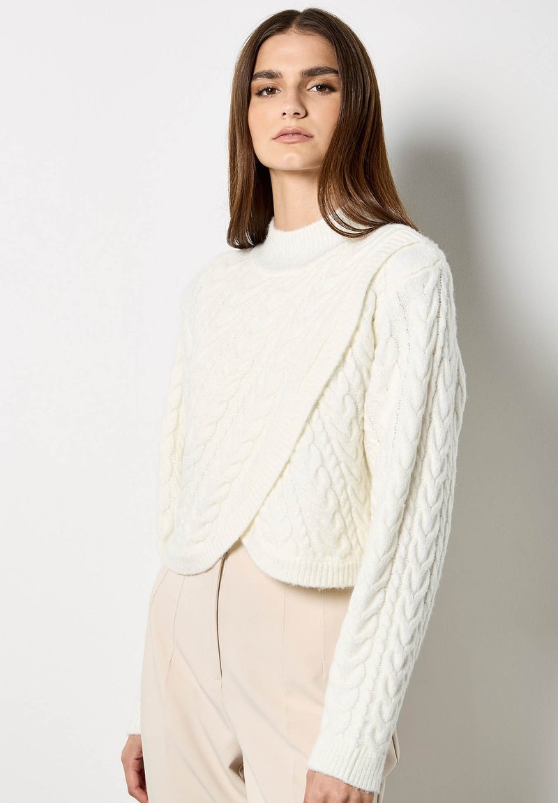 Maglione bianco a trecce con collo alto e design asimmetrico. Presenta un motivo a rilievo e maniche leggermente oversize.