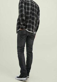 Jeans grises con bolsillos traseros, llevados con una camisa de cuadros en blanco y negro y zapatillas negras, vistos desde atrás sobre un fondo claro.