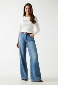 Witte lange mouwen top met grafische tekst, gecombineerd met wijde blauwe spijkerbroek. Bruine riem met zilveren gesp. Gladde textuur, casual stijl.