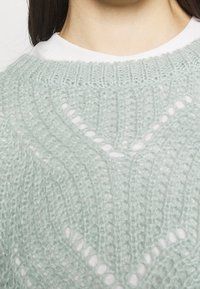 Pull en tricot vert clair avec un motif de tissage ouvert et un design en chevron texturé, porté sur un haut blanc, mettant en valeur un tissu doux et pelucheux.